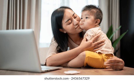 A happy Asian mom is playing and kissing her cute little son while working in her home office. family bonding, mom's life, happy motherhood - Powered by Shutterstock