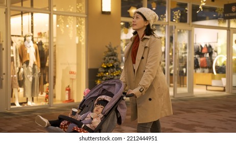 Happy Asian Mom Pushing A Baby Carriage And Doing Window Shopping For Christmas Presents On Downtown Street With Beautiful Light Decoration In The Evening.