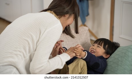 Happy Asian Mother And Baby Son Having Good Laugh While They Are Playing Tickle Together. On The Couch In The Living Room At Home.