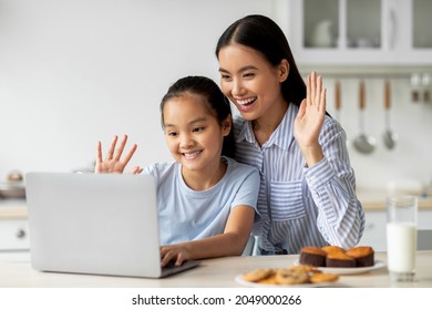 Happy Asian Mother And Daughter Having Video Call, Waving And Smiling At Laptop Screen, Sitting In Kitchen Interior, Copy Space. Pretty Girl And Her Mom Having Video Chat With Dad