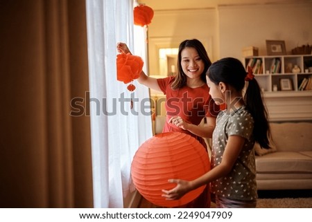 Happy Asian mother enjoying while hanging red lanterns with her daughter for Chinese New Year celebration at home.