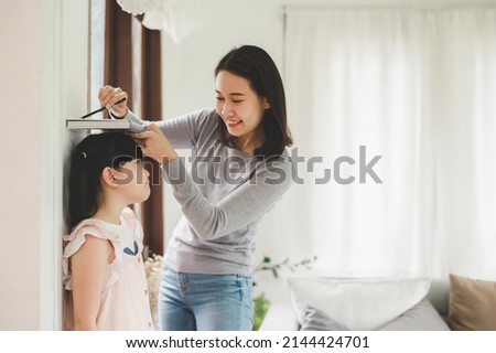 Happy Asian mother using book and pencil to measure height of little girl near wall at home
