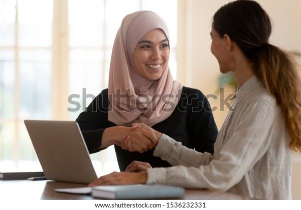 Happy asian muslim businesswoman sales manager\
shake hand of caucasian lady client make deal with female customer\
at meeting with laptop, diverse women partnership, respect and\
collaboration concept