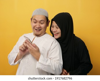 Happy Asian muslim husband shows phone to her wife, couple smiling looking at smartphone, aganst yellow background - Powered by Shutterstock