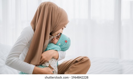 Happy Asian Muslim Mother Holding And Kissing Adorable Little Baby Daughter Wearing Hijab In Her Arms On White Bed In Bedroom. Arab Young Mom And Baby In Muslim Dress. Love Of Mother And Baby Concept.