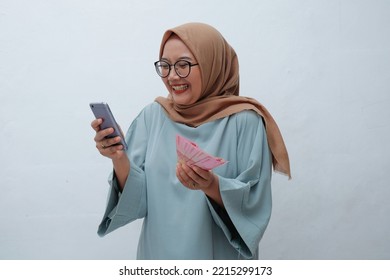 Happy Asian Muslim woman holding phone and cash money in Indonesian rupiah - Shutterstock ID 2215299173