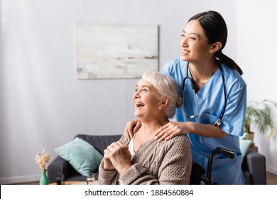 happy asian nurse touching shoulders of aged handicapped woman laughing in wheelchair