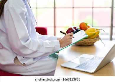 Happy Asian Nutritionist Working In Her Office In Hospital. Specialist In Nutrition Chatting To A Patient By Using Laptop Computer In The Office. Professional Nutrition Consultant Portrait.