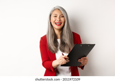 Happy asian office employee, woman in red blazer, working and laughing, holding clipboard with documents, standing over white background