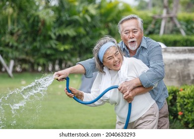 Happy Asian Old Couple Watering The Plants  In The Front Lawn At Home. Senior Man And Elder Woman Spend Time Together In Backyard . Mature Husband And Wife Lifestyle In Garden
