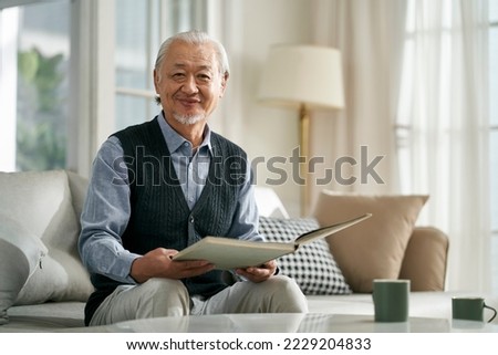 happy asian old man sitting on couch at home reading a book looking at camera smiling Stock photo © 
