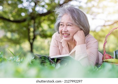 Happy Asian Old Senior Woman And Wear A Health Watch And Reading Book And Lying On The Picnic Mat In Park And Basket Of Fruit Besides. Concept Of Happy Elderly Woman After Retirement And Good Health