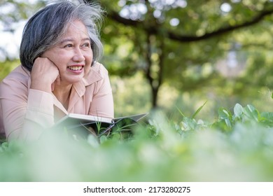 Happy Asian Old Senior Woman And Wear A Health Watch And Reading Book And Lying On The Picnic Mat In Park And Basket Of Fruit Besides. Concept Of Happy Elderly Woman After Retirement And Good Health