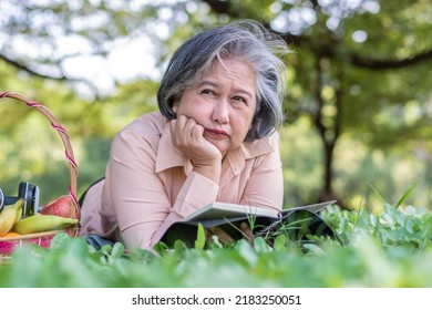 Happy Asian Old Senior Woman And Wear A Health Watch And Reading Book And Lying On The Picnic Mat In Park And Basket Of Fruit Besides. Concept Of Happy Elderly Woman After Retirement And Good Health