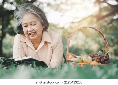 Happy Asian Old Senior Woman And Wear A Health Watch And Reading Book And Lying On The Picnic Mat In Park And Basket Of Fruit Besides. Concept Of Happy Elderly Woman After Retirement And Good Health