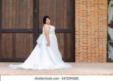 Happy Asian overweight model in beautiful bridal dress or white race wedding dress in many movement, healthy plump big size Asian model in elegant wedding dress walking outdoor at rustic brick wall 