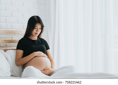 Happy Asian pregnant woman touching her belly with care in bedroom. She canâ??t wait to be mom.