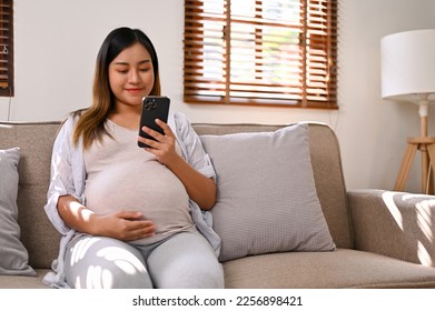 Happy Asian pregnant woman using her phone, reading an online column about motherhood preparation while relaxing on sofa in her living room. motherhood, pregnancy lifestyle  - Powered by Shutterstock