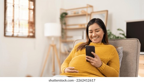 Happy Asian pregnant woman using her phone while relaxing on sofa in her living room - Powered by Shutterstock