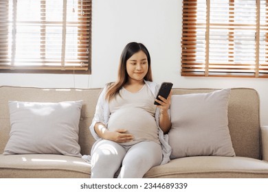 Happy Asian pregnant woman using smart phone while relaxing on sofa in her living room - Powered by Shutterstock