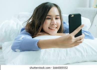 happy asian pretty woman in casual clothing smiling take selfie and video call on mobile phone and lying with pillow on bed in bedroom at home, lifestyle, internet technology, social network concept