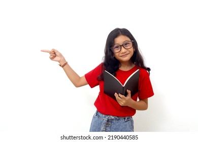 Happy asian schoolgirl standing while holding a book and pointing sideways. Isolated on white