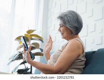 Happy Asian Senior Casual Woman Sitting On Couch, Waving Greeting At Digital Tablet Screen In Hand In White Room. Elderly Female Meet With Family By Video Call At Home. Older People With Technology.