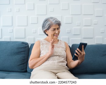 Happy Asian Senior Casual Woman Sitting On Couch, Waving Greeting At Digital Tablet Screen In Hand In White Room. Elderly Female Meet With Family By Video Call At Home. Older People With Technology.