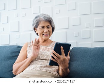 Happy Asian Senior Casual Woman Sitting On Couch, Waving Greeting At Digital Tablet Screen In Hand In White Room. Elderly Female Meet With Family By Video Call At Home. Older People With Technology.