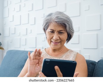 Happy Asian Senior Casual Woman Sitting On Couch, Waving Greeting At Digital Tablet Screen In Hand In White Room. Elderly Female Meet With Family By Video Call At Home. Older People With Technology.