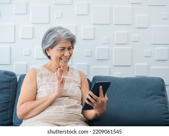 Happy Asian Senior Casual Woman Sitting On Couch, Waving Greeting At Digital Tablet Screen In Hand In White Room. Elderly Female Meet With Family By Video Call At Home. Older People With Technology.