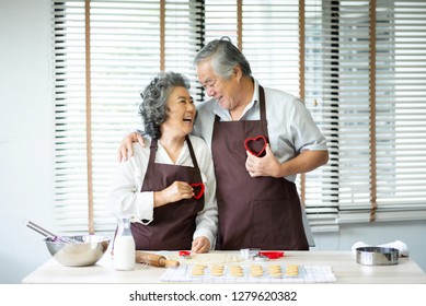 Happy Asian Senior Couple In Brown Aprons Are Holding Red Cookies Cutters In Heart Shape Over Their Chest. Looking Each Other. Having Fun.