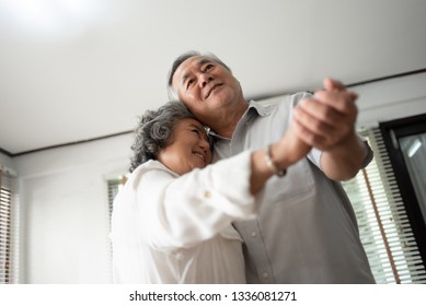 Happy Asian Senior Couple Dancing And Smiling At Home. Celebrating Wedding Anniversary.