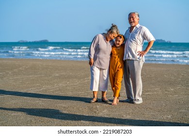 Happy Asian Senior Couple Family On Summer Travel Beach Vacation. Grandfather And Grandmother With Grandchild Girl Enjoy Outdoor Lifestyle Walking And Playing Together On The Beach At Summer Sunset.