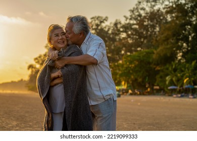 Happy Asian senior couple family relax and enjoy outdoor lifestyle travel on summer beach vacation. Elderly retired man cover his wife with a blanket and hugging together on the beach at summer sunset