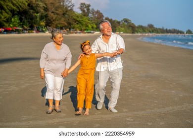 Happy Asian Senior Couple Family On Summer Travel Beach Vacation. Grandfather And Grandmother With Grandchild Girl Enjoy Outdoor Lifestyle Walking And Playing Together On The Beach At Summer Sunset.