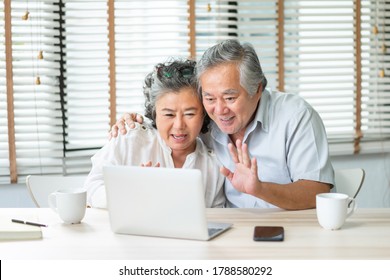 Happy Asian Senior Couple Making Video Chat And Waving Hands To Their Family On Laptop At Home. Smiling Elderly Man And Woman Enjoy With Internet Technology On Notebook Together, Mature Lifestyle