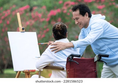 Happy Asian senior couple painting together on frame at park, blank frame, woman sitting and painting on wheelchair, blurred pink flowers on the background - Powered by Shutterstock