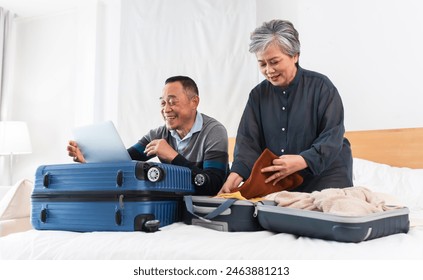 Happy Asian senior couple preparing for journey. Old man using laptop to planning and woman packing suitcases with excitement for trip, smiling, sitting on bed at home, getting ready vacation together - Powered by Shutterstock