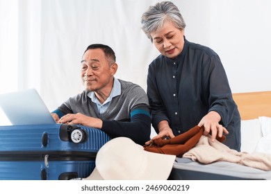 Happy Asian senior couple preparing for journey. Old man using laptop to planning and woman packing suitcases with excitement for trip, smiling, sitting on bed at home, getting ready vacation together - Powered by Shutterstock