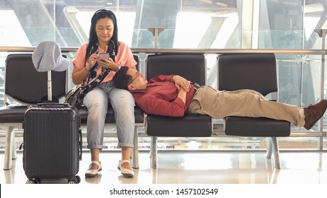 Happy Asian Senior Couple Sitting On Bench Using Smart Phone Sleeping Waiting For Departure At  Terminal Airport . Elderly Journey. Older Lovers . Travel. Flight. Retired . Together. Vacation