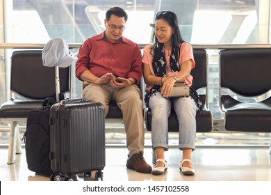 Happy Asian Senior Couple Sitting On Bench Using Smart Phone Waiting For Departure At  Terminal Airport . Elderly Journey. Older Lovers  Wife And Husband . Travel. Flight. Retired . Together. Vacation