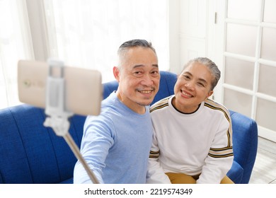 Happy Asian senior couple smile and take selfies photos and videos with smart phone and selfie stick together while sitting on a sofa in the living room. - Powered by Shutterstock