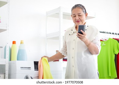 Happy Asian senior elderly old woman housewife using mobile phone while doing laundry at laundry room, grandma using smartphone while doing housework and cleaning clothes at house.