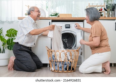 Happy Asian Senior Older Man Helping His Wife Put Clothes In Washing Machine In Laundry Room