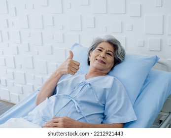 Happy Asian Senior Woman Patient Grey Hair Smiling, Thumb Up And Looking At Camera While Lying On Bed In Hospital. An Old Lady In Blue Patient Gown Is Sick On The Bed In Modern White Room.