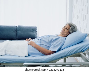 Happy Asian Senior Woman Patient Grey Hair Smiling And Looking At Camera While Lying On Bed In Hospital With Copy Space. An Old Lady In Blue Patient Gown Is Sick On The Bed In Modern White Room.