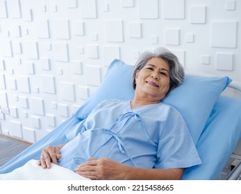 Happy Asian Senior Woman Patient Grey Hair Smiling And Looking At Camera While Lying On Bed In Hospital With Copy Space. An Old Lady In Blue Patient Gown Is Sick On The Bed In Modern White Room.