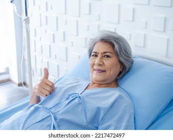 Happy Asian Senior Woman Patient Grey Hair Smiling, Thumb Up And Looking At Camera While Lying On Bed In Hospital. An Old Lady In Blue Patient Gown Is Sick On The Bed In Modern White Room.