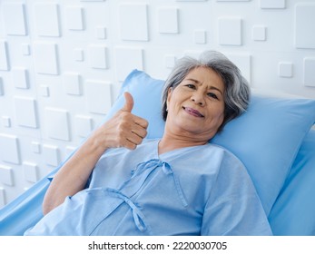 Happy Asian Senior Woman Patient Grey Hair Smiling, Thumb Up And Looking At Camera While Lying On Bed In Hospital. An Old Lady In Blue Patient Gown Is Sick On The Bed In Modern White Room.